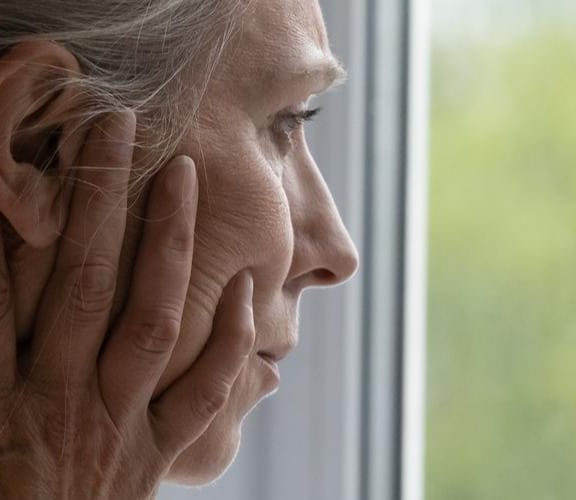 Elderly woman looking out a window with a pensive look on her face